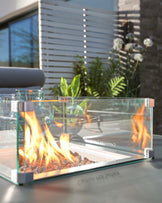 Modern outdoor glass fire pit table with visible flames and reflective stainless steel accents, set against an exterior backdrop with greenery.