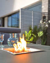 Modern outdoor patio table with a built-in fire pit feature, set against an architectural backdrop with blurred greenery, sunlight, and a partial view of a comfortable chair.