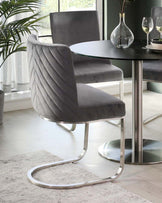 A modern dining chair with a grey velvet upholstered seat and backrest, featuring vertical channel tufting and a unique, sleek cantilever base in a polished chrome finish. Paired with a round dining table with a black tabletop and a central chrome pedestal base. The setting is complemented by minimalist decor, including a clear glass vase and wine glasses atop the table.