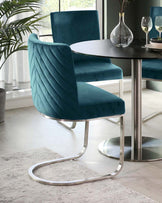 Elegant contemporary dining set featuring a round, black tabletop with a sleek metallic pedestal base and a pair of luxurious teal blue velvet chairs with unique geometric quilted stitching, complemented by smooth, curved chrome cantilever bases.