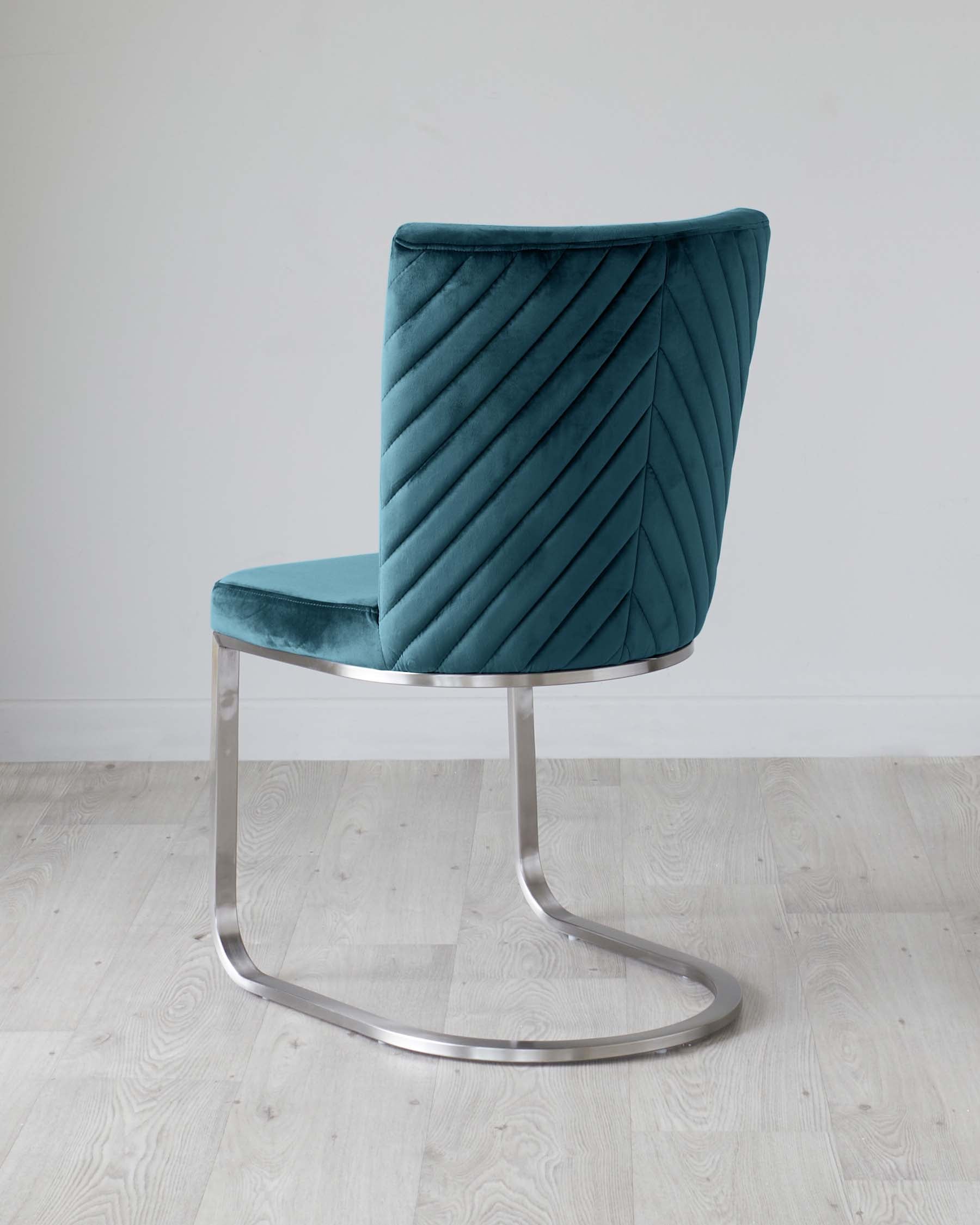 Modern teal velvet accent chair with a curved, channel-tufted backrest and a smooth, flat seat cushion. The chair is supported by a sleek, chrome-finished, cantilevered metal base, set on a light wooden floor against a white background.