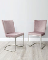 A pair of modern minimalist dining chairs with blush pink upholstery and sleek, cantilevered silver metal frames on a light wooden floor against a white wall.