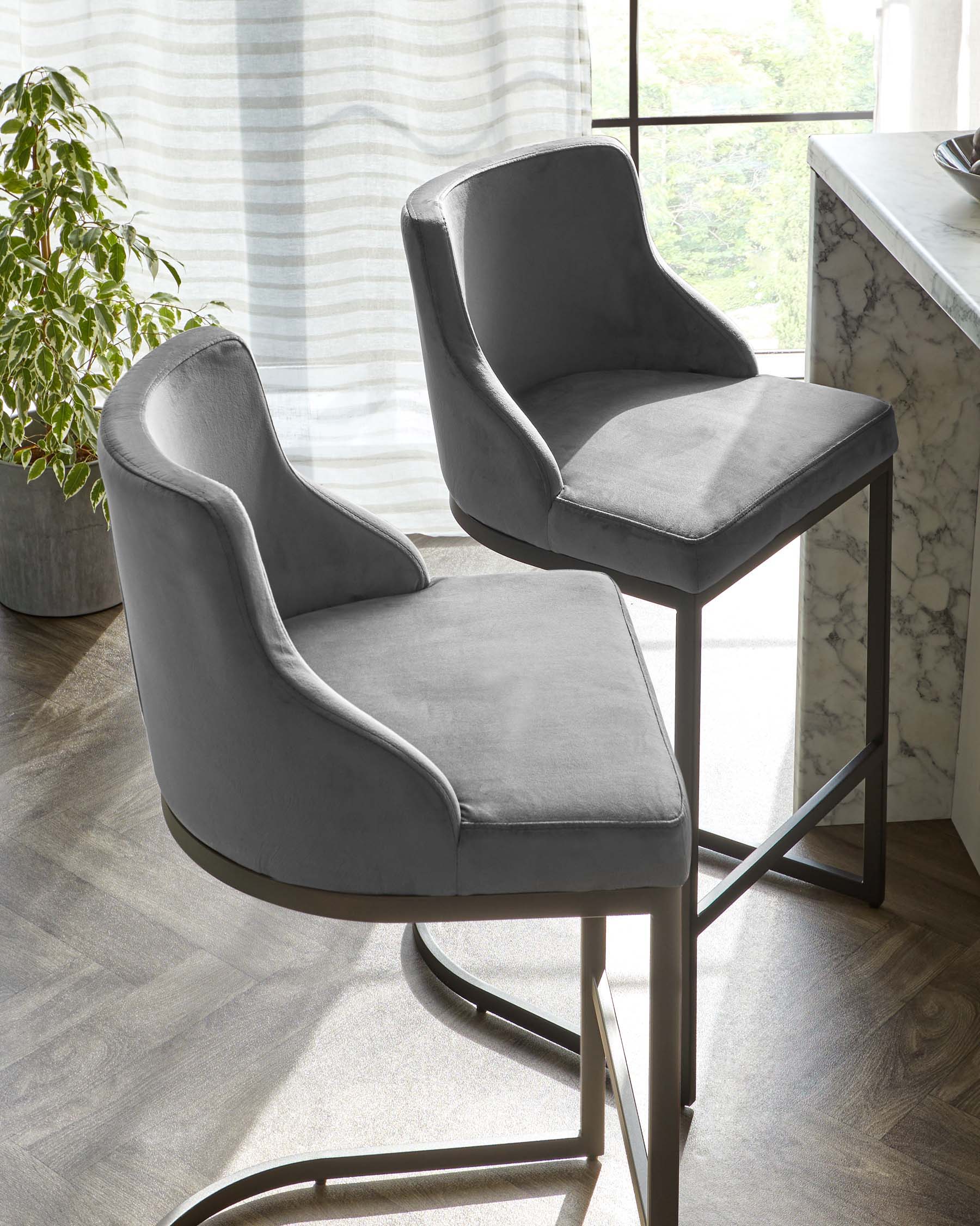 Two contemporary grey upholstered dining chairs with a smooth fabric finish and curved backrests, featuring sleek black metal legs, displayed in a modern room with a marble countertop and a light curtain backdrop.