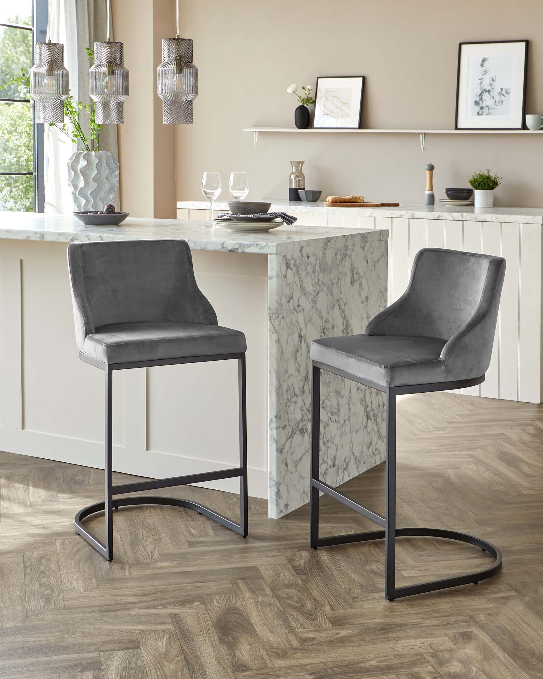 Elegant modern kitchen with two grey velvet upholstered bar stools featuring sleek black metal legs. The bar stools are positioned at a white marble countertop island that contrasts with light wood flooring.