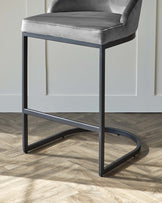 Modern grey velvet bar stool with a sleek, black metal frame against a white wall on a herringbone wood floor.