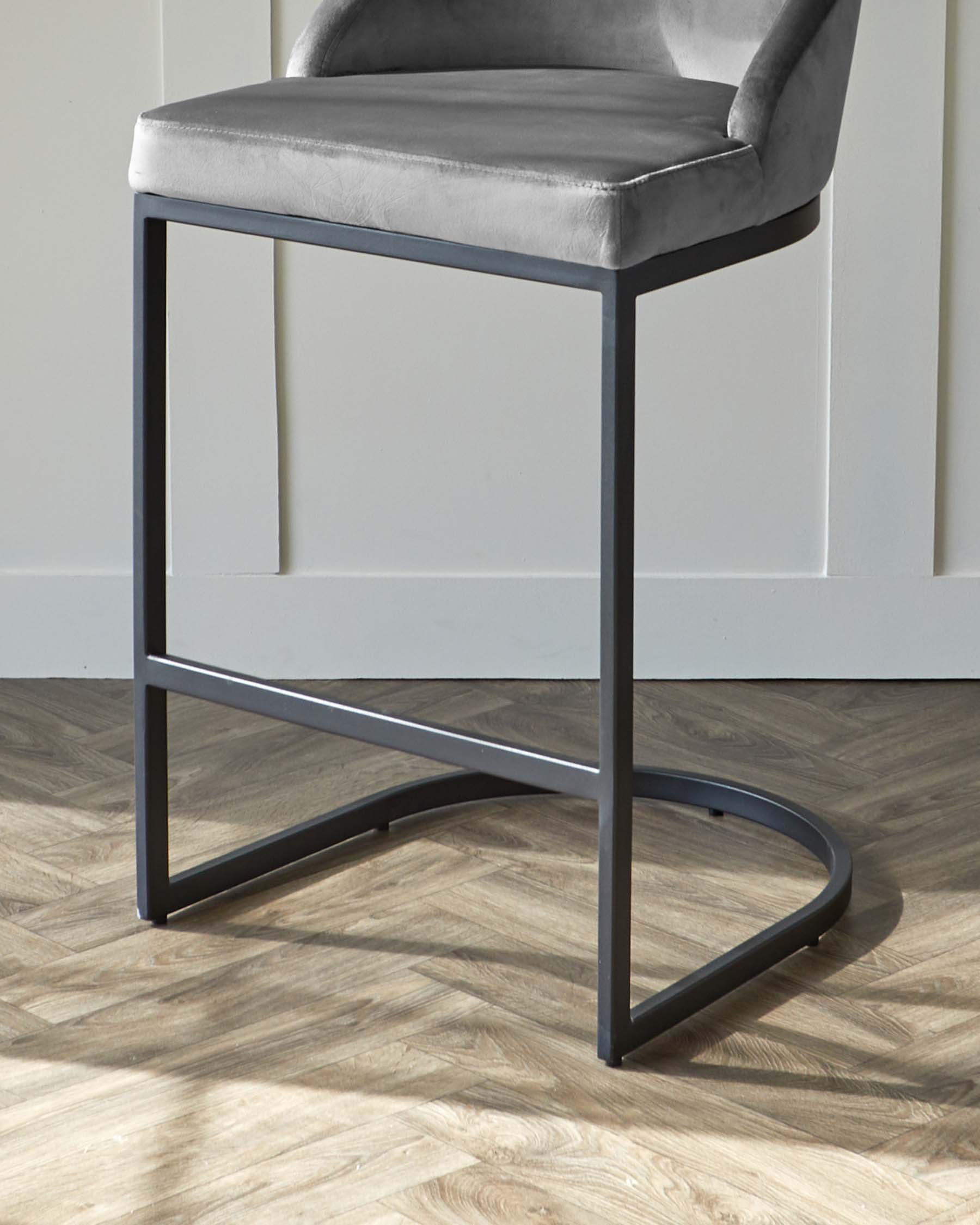 Modern grey velvet bar stool with a sleek, black metal frame against a white wall on a herringbone wood floor.