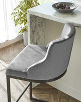 Elegant modern dining chair with a high back and armrests, upholstered in grey fabric with a subtle sheen. The chair features a smooth, curved silhouette and is paired with a sleek black metal frame. To the side, a glimpse of a white marble-top kitchen island or table with a decorative bowl of fruit is visible, complementing the luxurious feel of the scene.