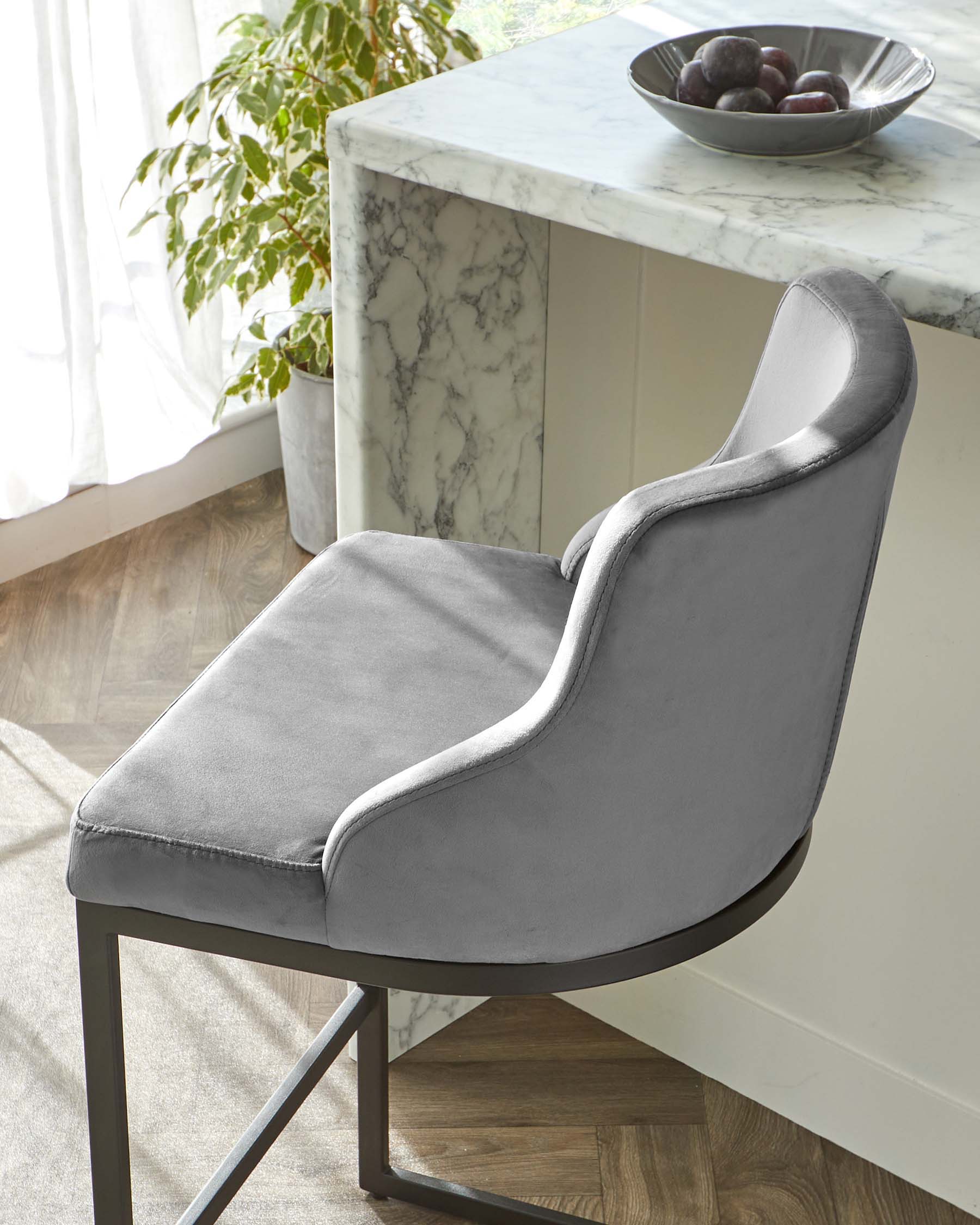 Elegant modern dining chair with a high back and armrests, upholstered in grey fabric with a subtle sheen. The chair features a smooth, curved silhouette and is paired with a sleek black metal frame. To the side, a glimpse of a white marble-top kitchen island or table with a decorative bowl of fruit is visible, complementing the luxurious feel of the scene.