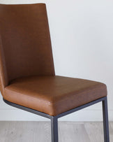 Modern dining chair with a sleek, minimalist design featuring a smooth, tan faux leather upholstery and a sturdy, matte black metal frame, set against a plain white background with a light wooden floor.