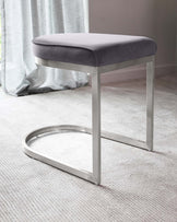 Modern minimalist stool with a grey upholstered seat and sleek, polished chrome frame featuring a unique circular base design. Positioned on a textured light beige carpet with a sheer curtain backdrop.