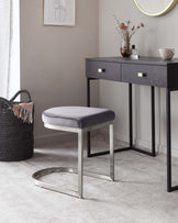 Modern minimalist furniture set featuring a sleek black console table with two drawers accented with brass handles, and a complementing grey upholstered vanity stool with a polished silver base.