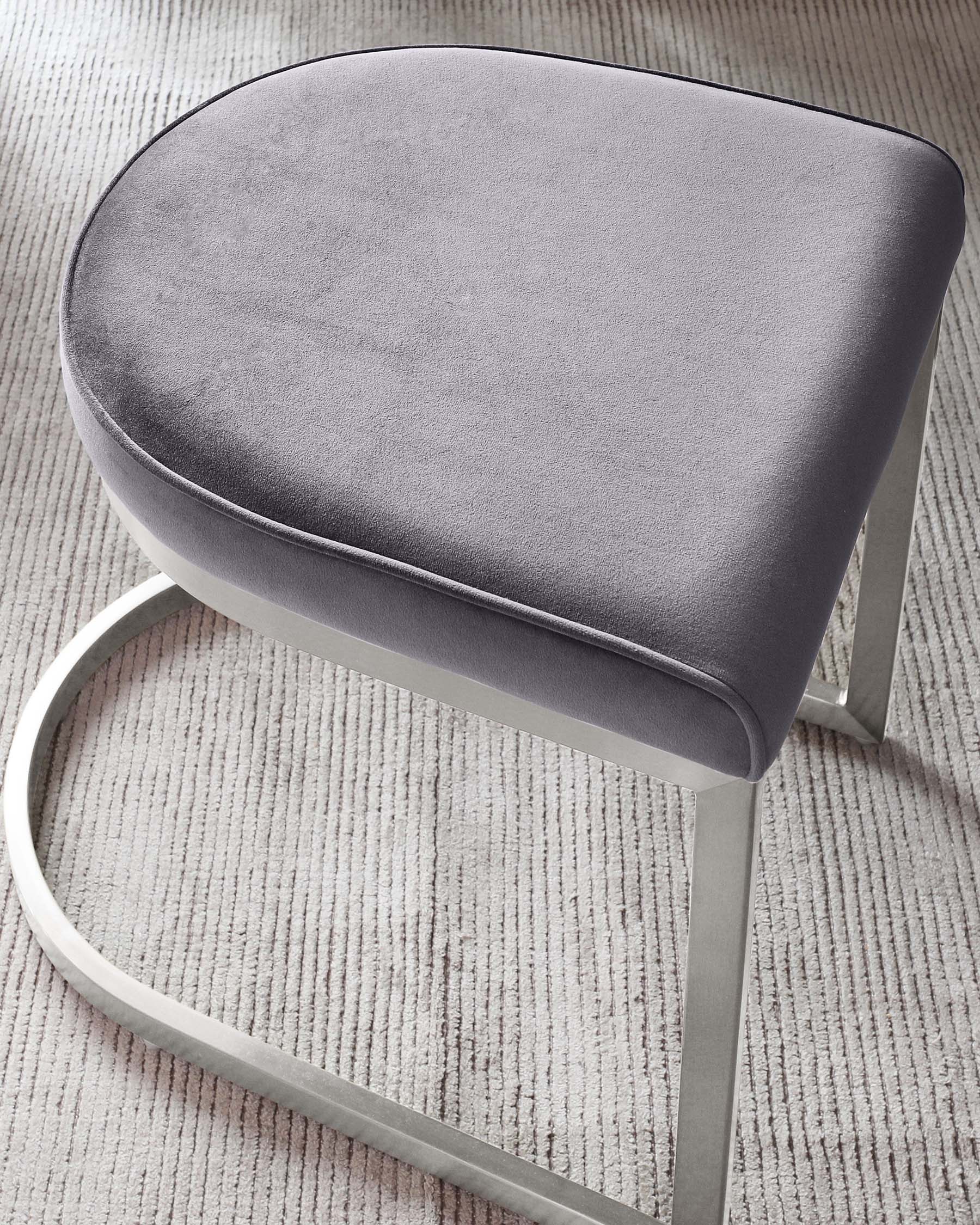 Modern grey fabric upholstered stool with a sleek, curved seat and sturdy metallic silver frame, showcased on a textured carpet.