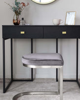 A modern, minimalist vanity table in black with sleek golden handles, paired with a round, padded stool upholstered in grey fabric, featuring a chic metallic base.