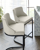 Two modern dining chairs with cream upholstery and black metal frames, featuring an elegant curved backrest and cushioned seat.