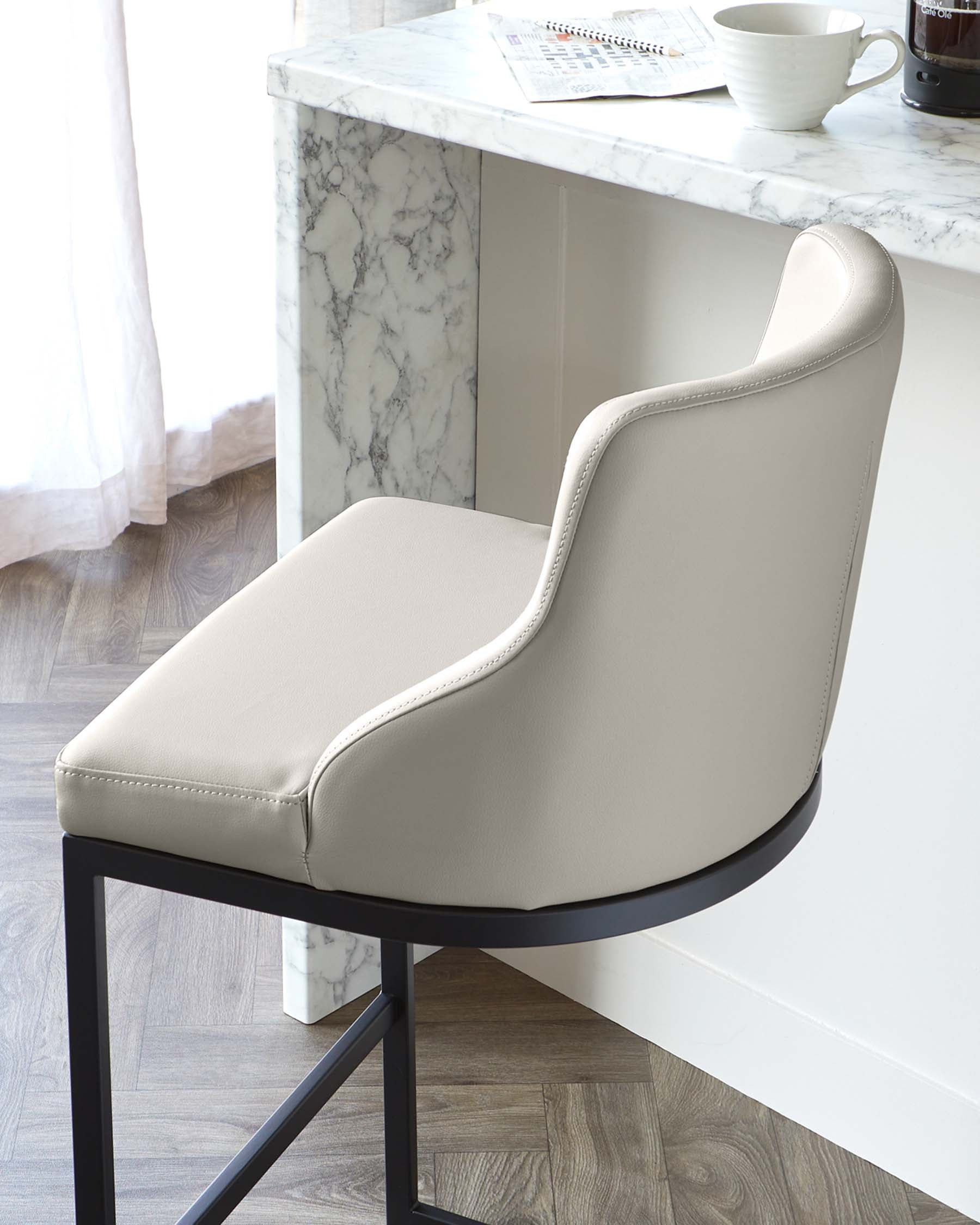 Elegant modern dining chair with a light beige upholstered seat and backrest featuring a distinctive curved design and subtle nail head trim details. The chair has a sleek black metal base providing sturdy support, set against a sophisticated marble countertop kitchen island.