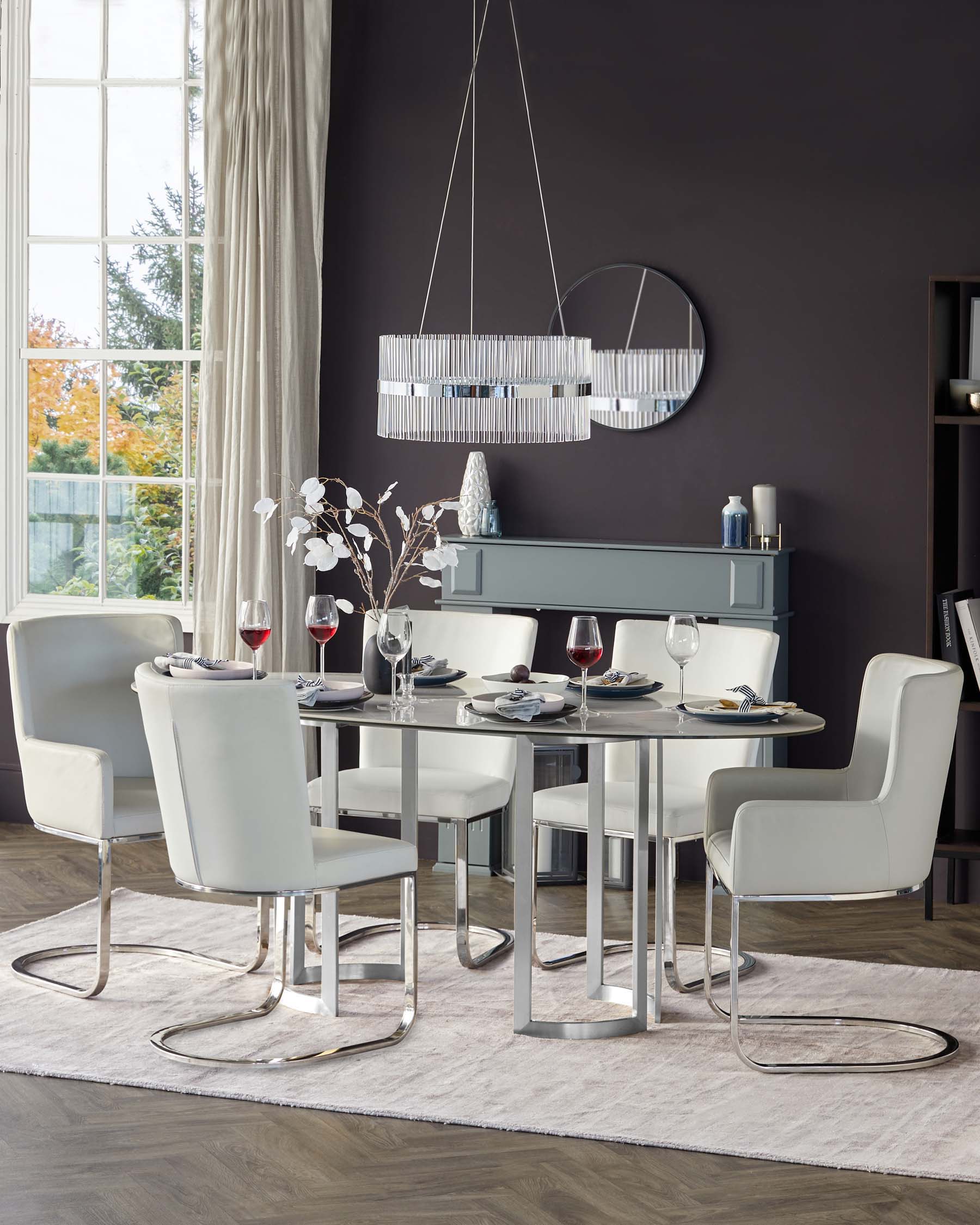 Modern dining room with a rectangular white marble table supported by a silver metal base, surrounded by six white leather upholstered chairs with curved backrests and sleek chrome cantilevered legs. A sideboard in a matching pale colour palette stands against the wall under a circular mirror, accented with decorative items and flanked by a tall, dark shelf. A contemporary chandelier with a horizontal design hangs above the table, complementing the stylish and elegant interior.