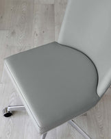 Modern light grey office chair with a sleek design, featuring a curved backrest and a flat seat cushion on a minimalist frame with metallic legs and black casters, displayed on a light wooden floor.