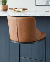 Elegant modern bar stool with a curved back, upholstered in caramel brown leather with visible stitching details, and supported by thin black metal legs.