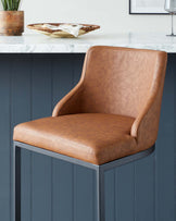 Modern dining chair with smooth cognac-coloured faux leather upholstery, featuring a curved backrest and cushioned seat resting on a clean-lined, dark metallic frame. The chair is displayed against a blue panelled wall next to a marble countertop accentuated with a wooden bowl and napkin, suggesting a contemporary kitchen setting.