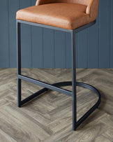 Modern bar stool with sleek black metal frame and camel-hued leather cushioned seat and backrest, featuring a unique X-base footrest design.