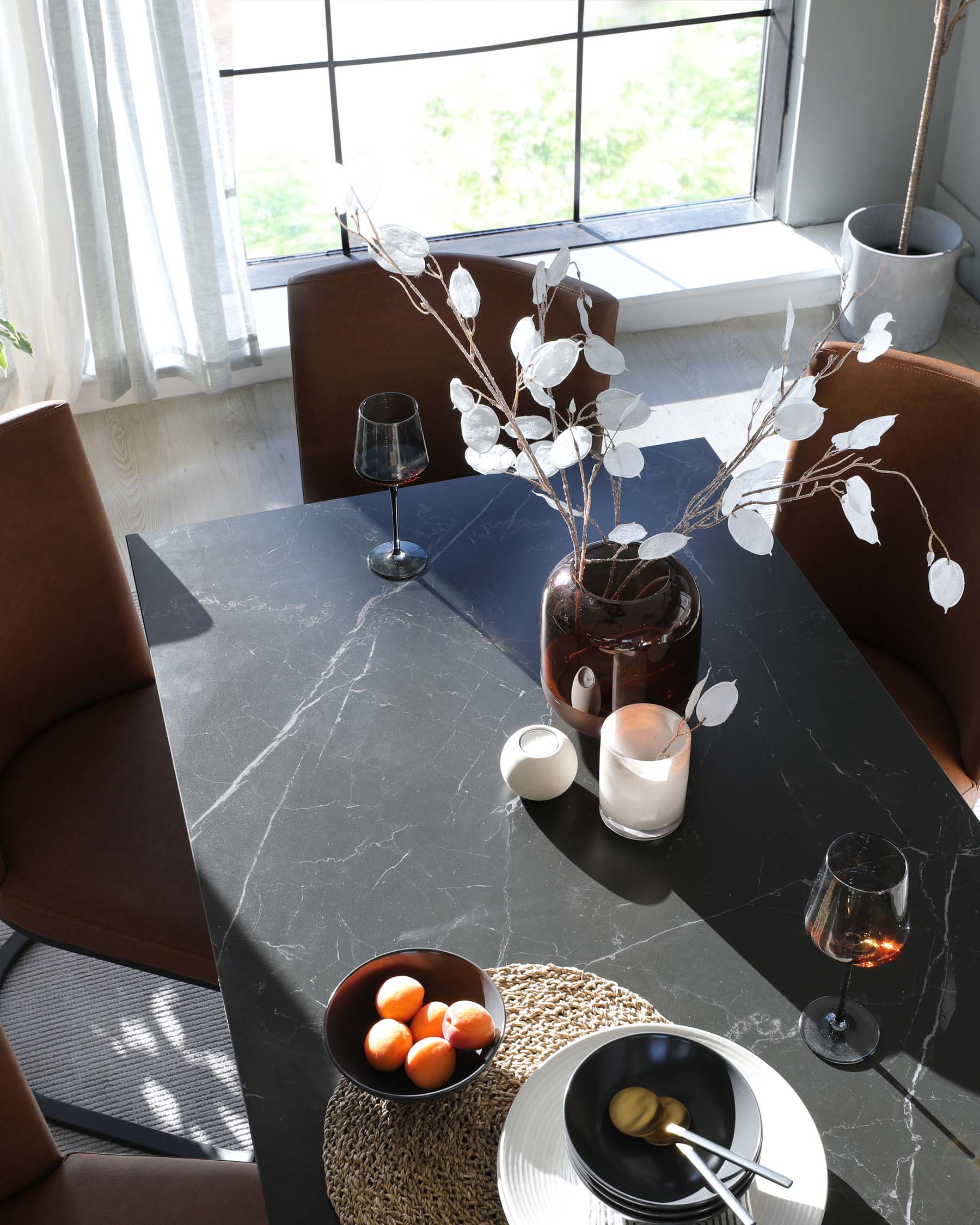 A modern rectangular dining table with a dark marble tabletop, accompanied by two curved, brown leather dining chairs with a seamless back and seat design. The table is set with a bowl of fruit, decorative candles, glasses, and plates atop a woven circular placemat. The scene is bathed in natural light from a large window with sheer curtains, creating a warm and inviting atmosphere.