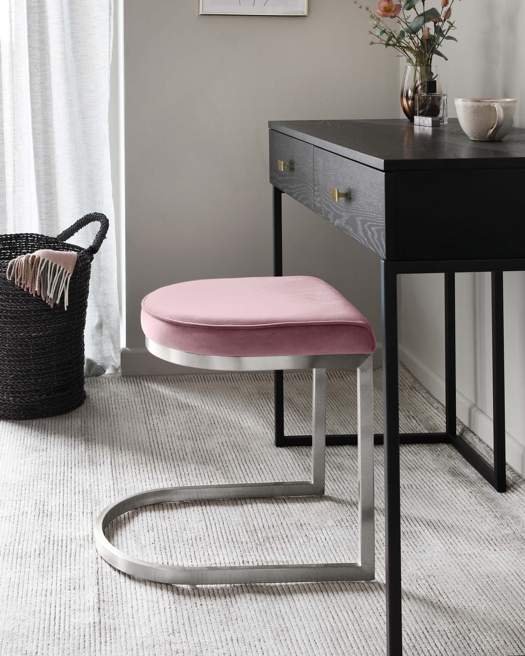 A modern black wooden console table with sleek lines features two drawers with brass handles, set against a light wall above a textured off-white rug. Accompanying the table is a contemporary round ottoman with a blush pink velvet cushion top and a unique, geometric, silver metal base that curves to the floor. The setting is complemented by soft natural light and a minimalist aesthetic.