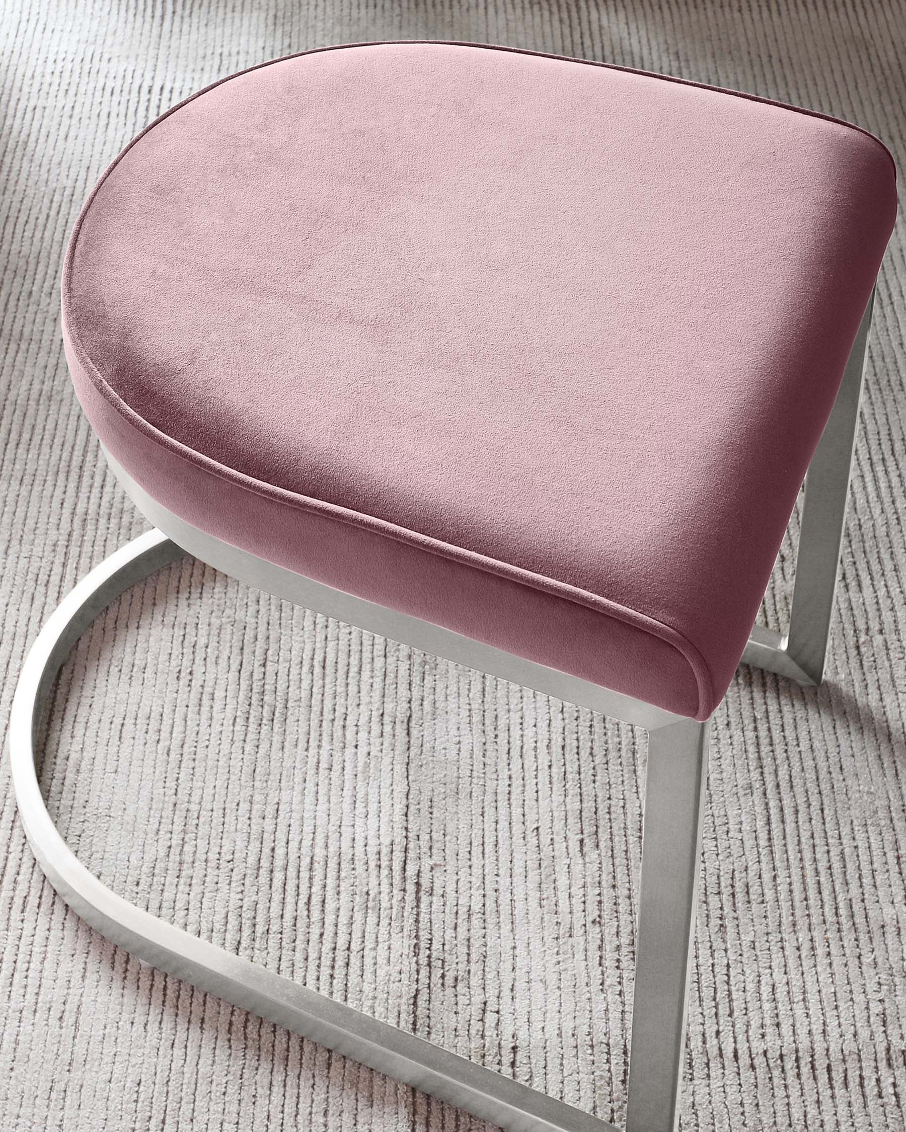 Modern blush pink velvet ottoman with a sleek silver metal base on a textured grey carpet.