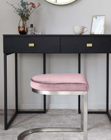 Elegant black console table with sleek metal legs and two drawers featuring brass handle accents, paired with a contemporary round-edged, blush-coloured upholstered stool with a metallic base.