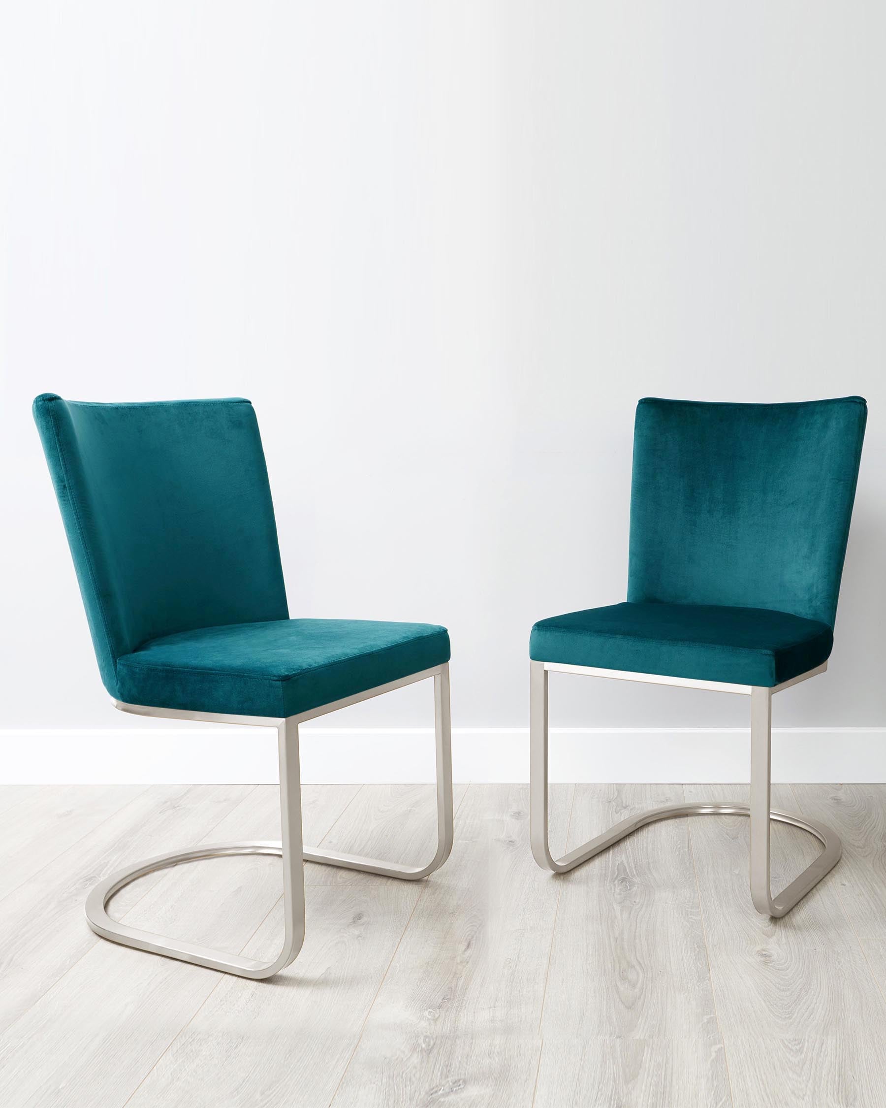 Two modern chairs with teal velvet upholstery and sleek metallic silver cantilever bases on a light hardwood floor against a white wall.