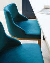 Elegant teal upholstered dining chairs with a smooth velvet finish and sleek golden metal legs placed beside a white minimalist dining table with architectural blueprints spread out on it, set against a backdrop of a light wood floor and a dark door frame.