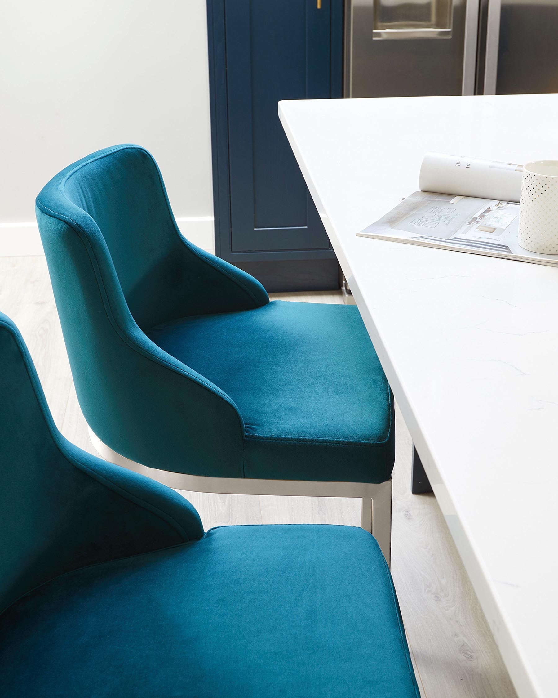 Elegant, modern dining chairs with a deep teal blue velvet upholstery and a sleek, curved backrest design, paired with a contemporary white dining table featuring clean lines and a glossy finish.