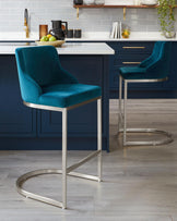 Two contemporary counter-height bar stools with plush teal velvet upholstery and sleek metallic silver frames in a stylish kitchen setting.