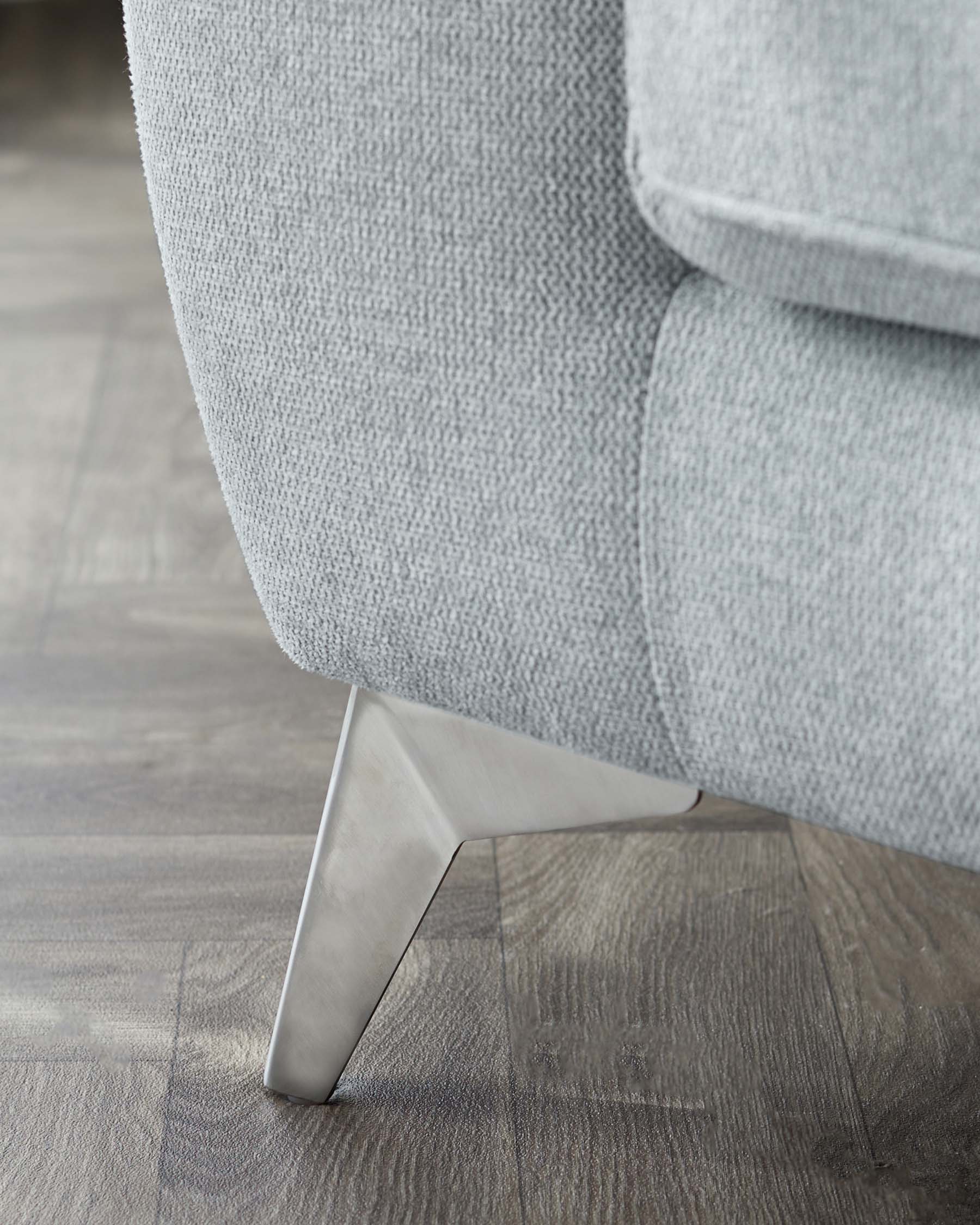 Close-up of a modern sofa's corner with light grey fabric upholstery and a sleek, angular metal leg on a wooden floor.