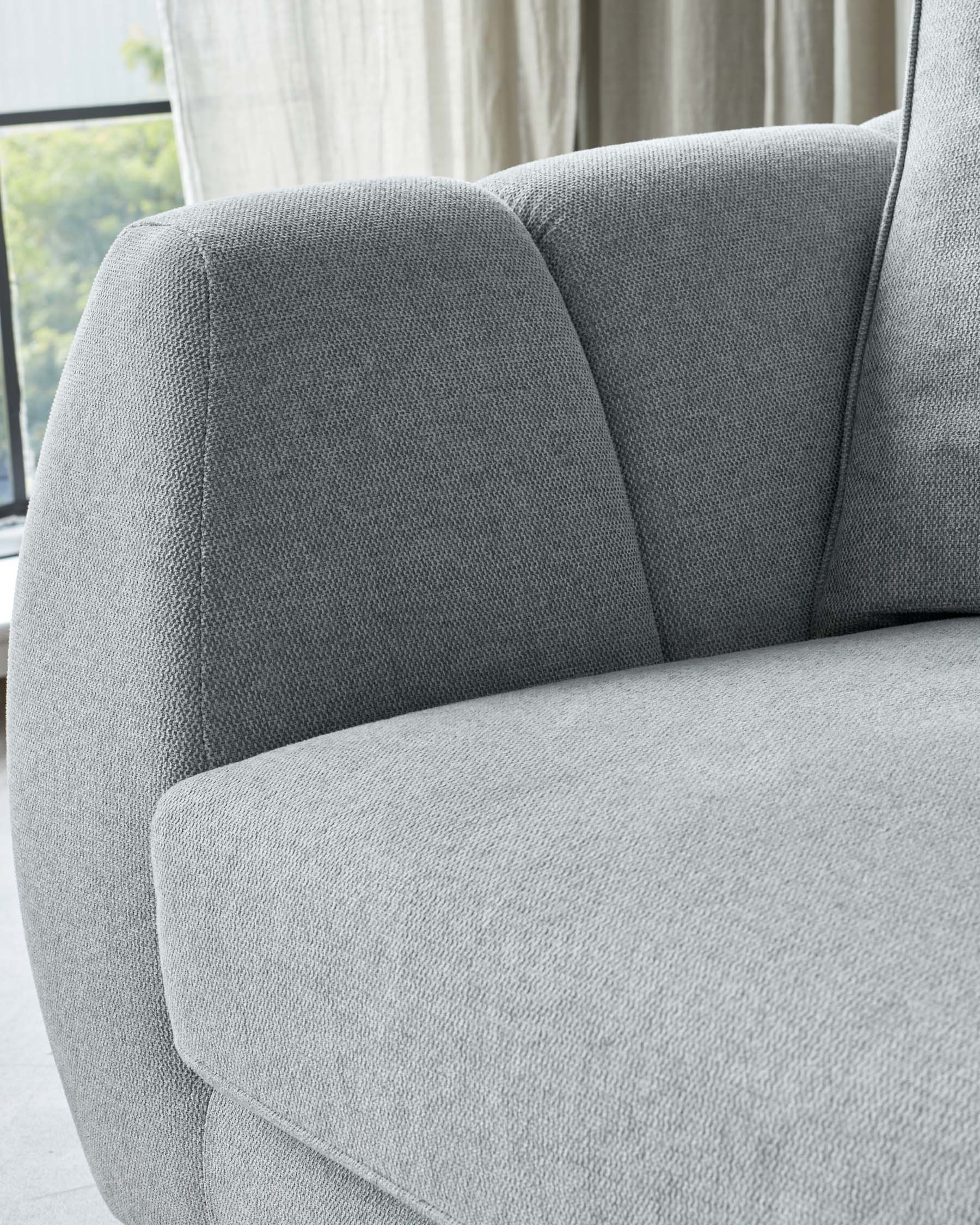 Close-up of a modern light grey fabric sofa with textured upholstery, showcasing its padded armrest, cushioned seat, and supportive backrest. The background features sheer curtains and a glimpse of greenery outside the window, indicating a bright and airy room setting.