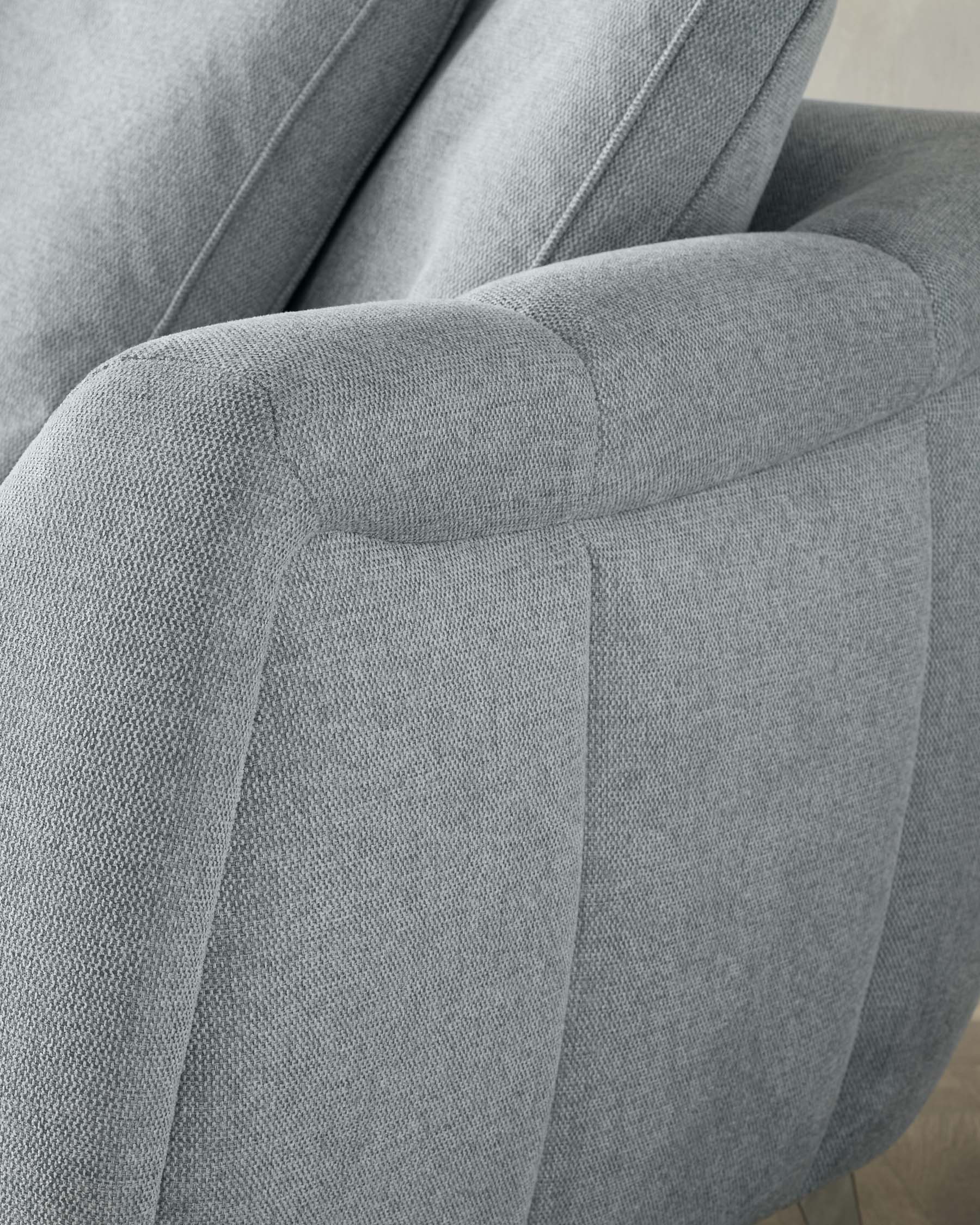 Close-up of a modern armchair with textured light grey upholstery. The chair features clean lines with a rounded armrest and stitched detailing, suggesting comfort and contemporary style.