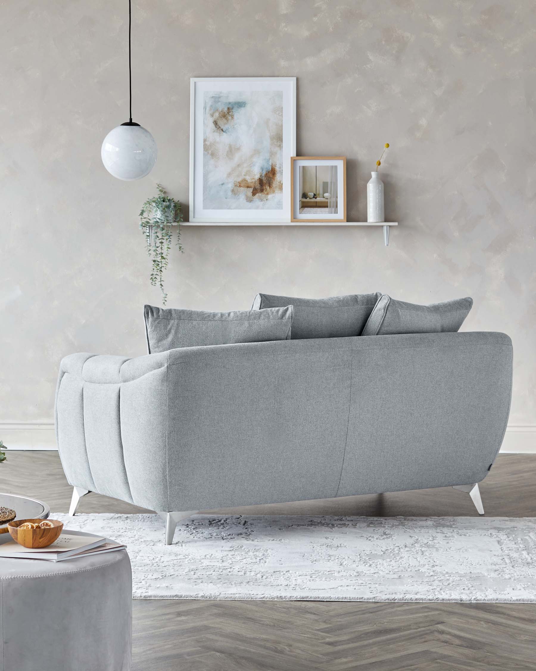 Modern light grey sofa with clean lines and tufted cushions, featuring tapered white legs, accompanied by a small, round grey ottoman with a smooth surface and matching white legs. A minimalist white floating shelf adorns the wall above, decorated with framed abstract art and a simple vase.