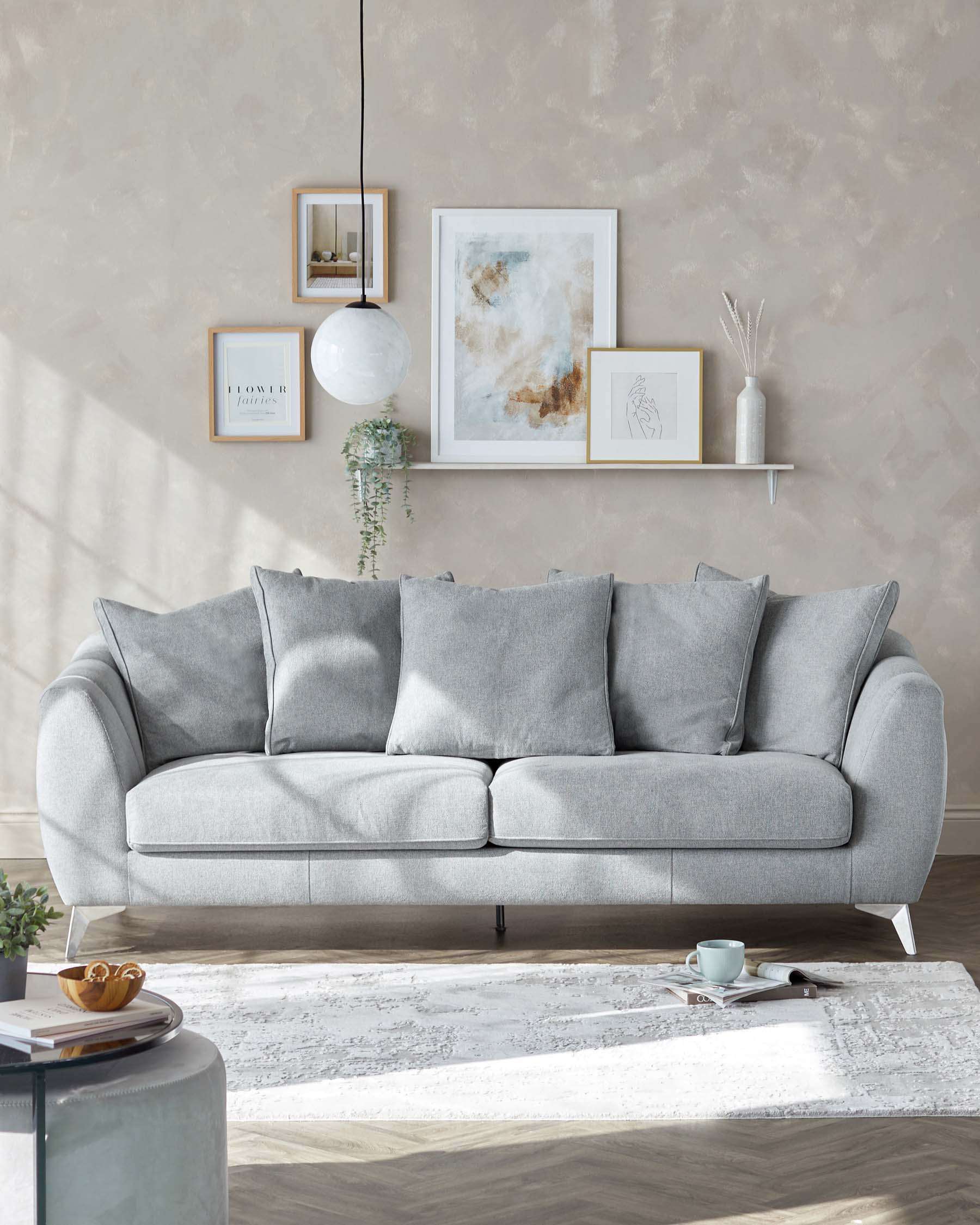 A modern light grey fabric sofa with three seat cushions and five back cushions, featuring a minimalist design with rounded edges and elegant metal legs. Positioned atop a textured off-white area rug, the sofa is complemented by a dark round side table and simple wall-mounted shelf adorned with decorative items.