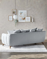 Elegant contemporary grey fabric sofa with tufted backrest and plush seat cushions, featuring rounded edges and angled wooden legs, complemented by a set of matching grey throw pillows. Positioned on a textured white and grey area rug over a wooden floor, the sofa is part of a stylish living room setting enhanced by a minimalistic white floating shelf with decorative items and framed artwork on the wall above.