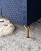 Contemporary navy blue sideboard with textured front panels and brass accent legs, complemented by light grey carpeting and a white faux fur accent rug. A clear glass tumbler and an open magazine rest beside the sideboard.