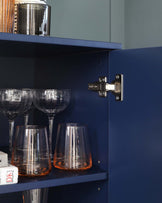 A deep blue, contemporary cabinet featuring a silver hinge on the door. The partially visible interior shelf holds elegant glassware, including upturned wine glasses and amber-bottomed tumblers. On top of the cabinet, a portion of a silver cocktail shaker with a striped design is visible. The sleek design and bold colour of the cabinet add a modern touch to any living space.