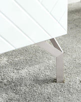 Close-up of a modern white cabinet with a geometric pattern on its surface and sleek, silver metal legs, placed on a textured grey shag rug.