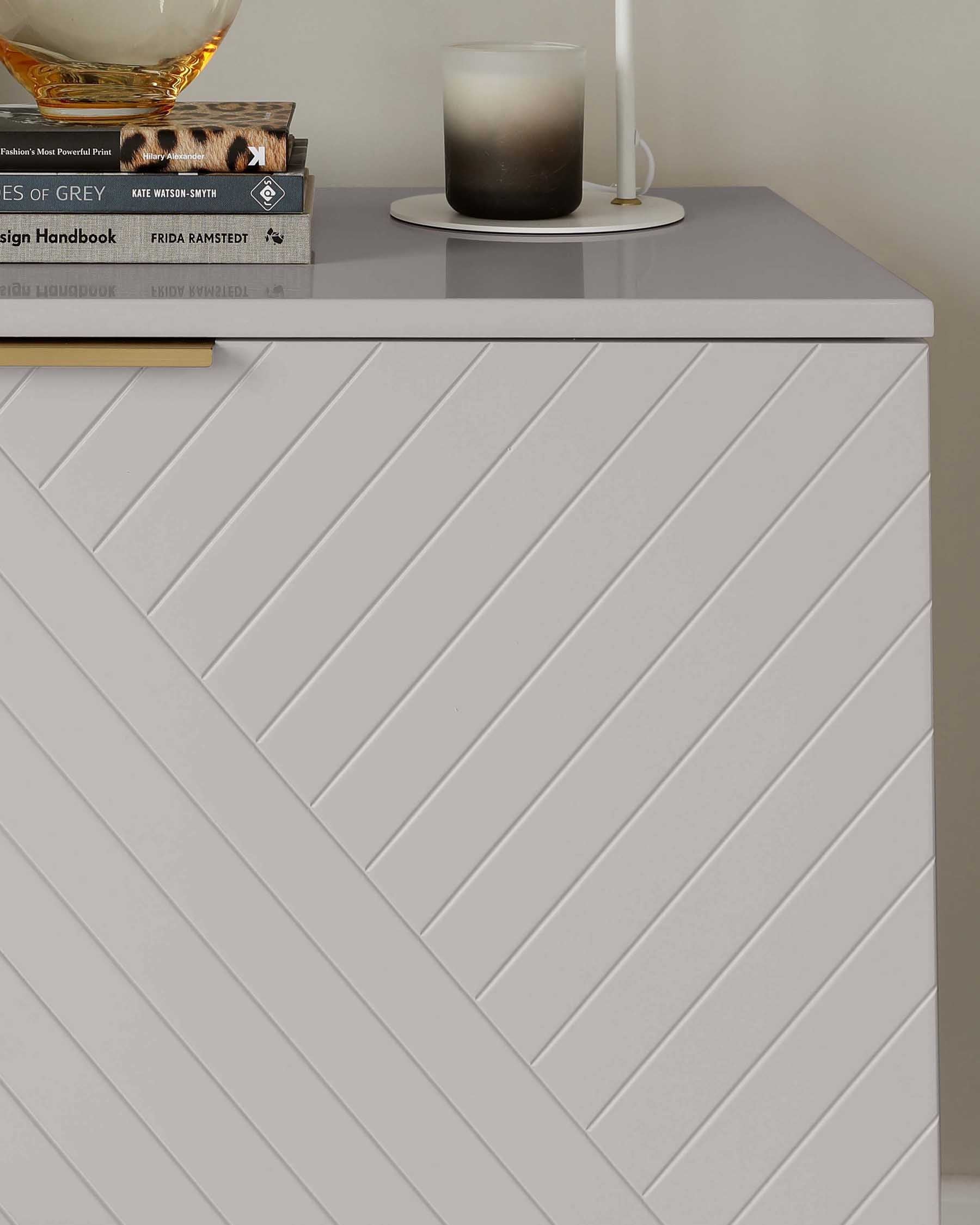 Modern minimalist white side cabinet with a chevron panel design and clean lines, topped with a stack of books, a scented candle, and a candle holder.