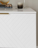 A modern white bedside table with diagonal groove design and a smooth flat top, displaying a stack of books, a decorative glass bowl, and a black candle on a plate.