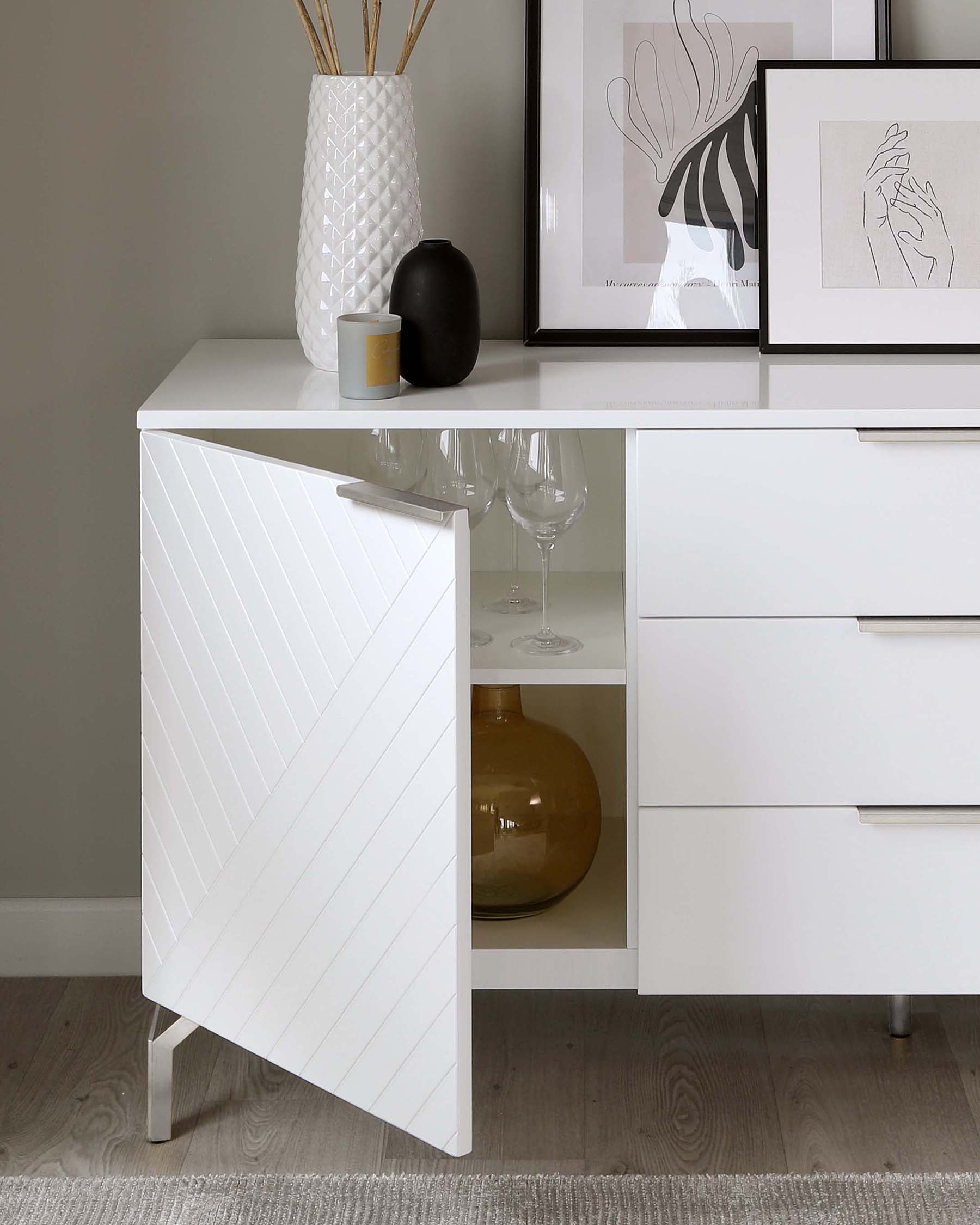 White modern sideboard with chevron patterned door, three drawers to the right, and an open shelf displaying a vase. Elegant, minimalistic design suitable for contemporary interiors.