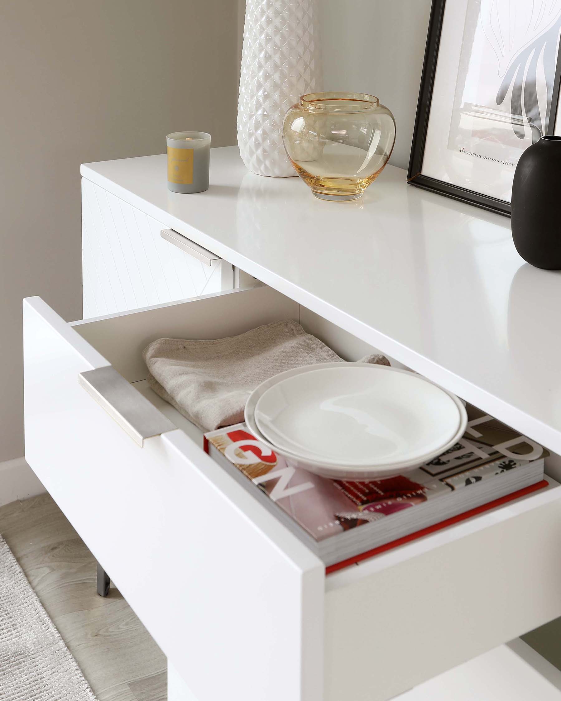 Modern white sideboard with a sleek design, featuring a partially opened drawer revealing a white ceramic dish, linen, and assorted magazines, accentuated by a minimalistic metal handle. The top of the sideboard is adorned with decorative items including a textured white vase, a clear glass vase with gold detailing, a small candle, and a framed artwork alongside a matt black vase, set against a neutral wall.