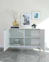 Modern minimalist white sideboard with gold accented legs featuring two open shelves and one closed compartment, partially open to reveal stacked dinnerware. The top surface is styled with decorative books, a vase with pampas grass, and a black decorative urn. Two framed artworks hang on the wall above the sideboard.