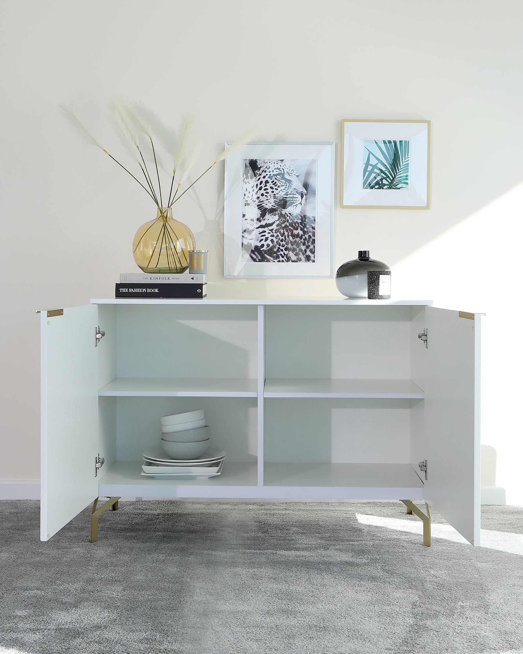 Modern minimalist white sideboard with gold accented legs featuring two open shelves and one closed compartment, partially open to reveal stacked dinnerware. The top surface is styled with decorative books, a vase with pampas grass, and a black decorative urn. Two framed artworks hang on the wall above the sideboard.