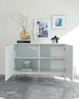 Modern white sideboard with clean lines and minimalist design. Features two levels of shelving with an asymmetrical partition, angular metal legs, and square-cut handles on swinging cabinet doors. The sideboard is accessorized with decorative items and flanked by framed wall art.