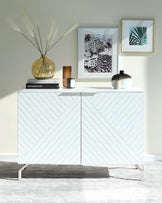 A contemporary white sideboard cabinet with a distinctive chevron pattern on the front and sleek, minimalist metal legs, positioned against a light wall on a textured grey area rug. The sideboard is accessorized with decorative items including a golden round vase with tall pampas grass, a stack of hardcover books, a small candle, and a stylish round black and gold vase. Two framed artworks hang on the wall above, one featuring a leopard and the other showcasing an abstract plant design.