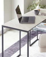 Modern minimalist-style office desk with a marble-patterned surface and a sleek black metal frame, accompanied by a cosy beige upholstered chair with a cushioned seat and no visible armrests. The setting includes a laptop, a ceramic mug, a notebook, a decorative vase with greenery, and designer books resting on a lower glass shelf, all atop a striped grey and white area rug.